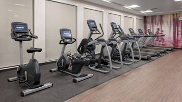 Fila de bicicletas estáticas y cintas de correr en un gimnasio con un espejo y un reloj en la pared.