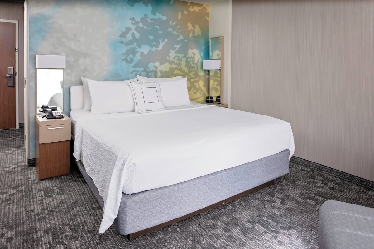 A hotel room with a large bed, two bedside tables, a nightstand, a wall-mounted lamp, and a door.