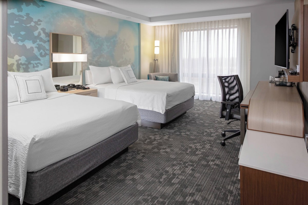 A hotel room with two beds, a desk, a chair, a TV, and a large window with curtains.