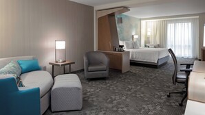 A hotel room with a sofa, armchair, ottoman, a desk with a lamp, a bed, and a large window with curtains. - Courtyard by Marriott Cincinnati Mason (Mason)