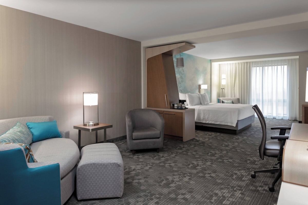 A hotel room with a sofa, armchair, ottoman, a desk with a lamp, a bed, and a large window with curtains.