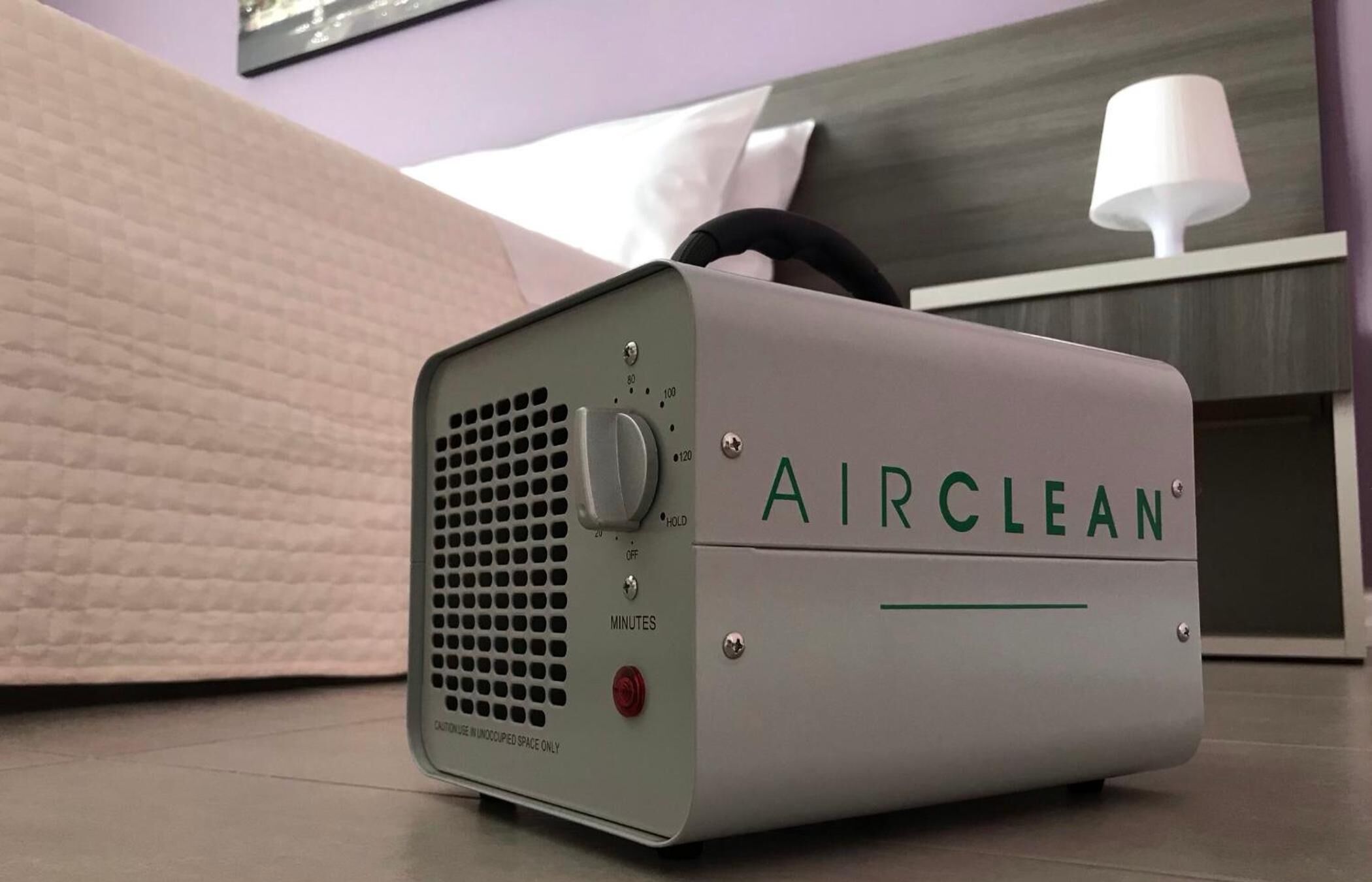 A portable air purifier labeled "AIRCLEAN" in a hotel room with a bed, pillows, and a nightstand with a lamp.