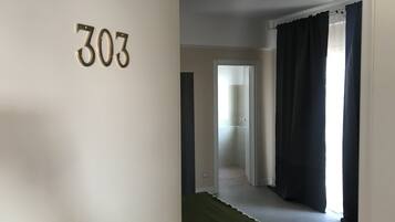 Una habitación con la puerta número 303 visible, un pasillo que conduce a otra habitación y una ventana con cortinas oscuras.
