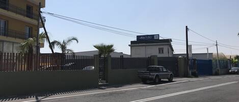 Una veduta di strada con un furgone bianco parcheggiato vicino a un'area recintata, un edificio con balconi e cielo sereno.