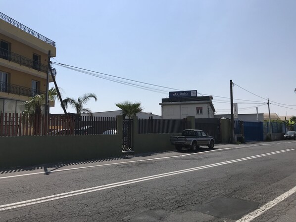 Vista da rua com uma caminhonete branca estacionada perto de uma área cercada, um prédio com sacadas e céu claro.
