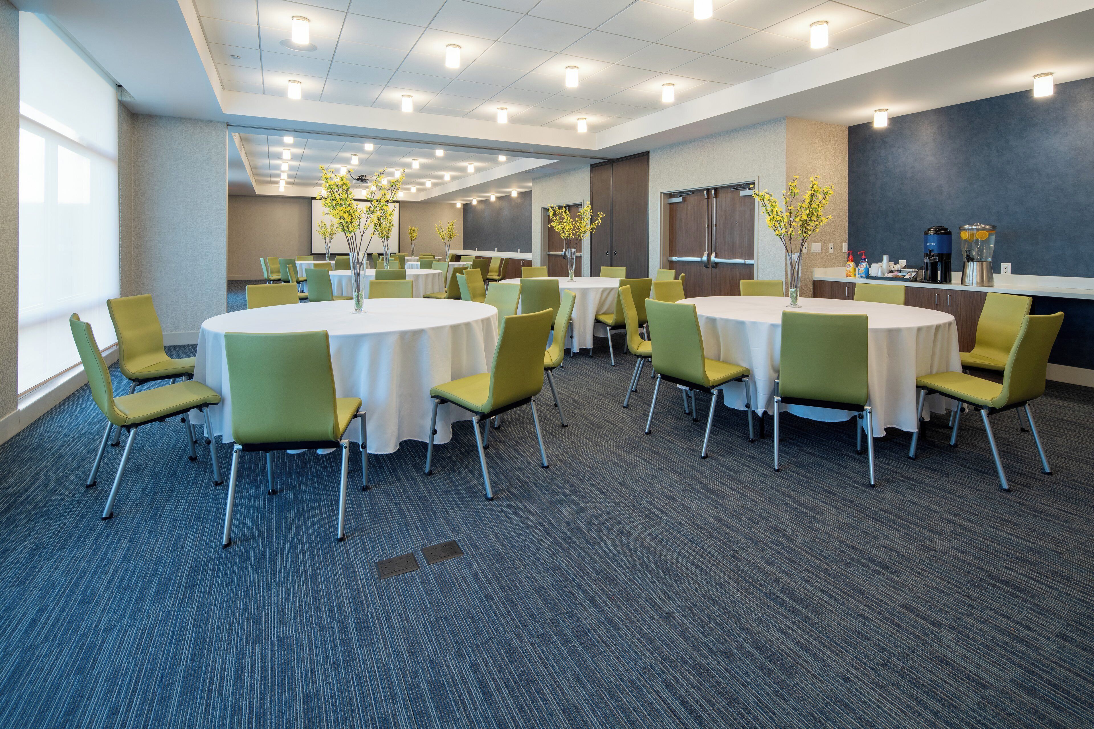 A conference room with multiple tables and chairs, equipped with ceiling lights and a projector screen.