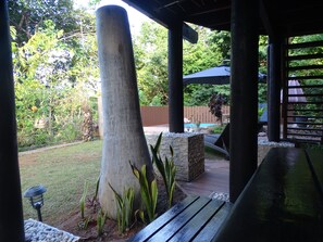 A covered outdoor area with a stone pillar, a wooden bench, and a view of a swimming pool and garden. - Gecko Villa (Praslin Island)