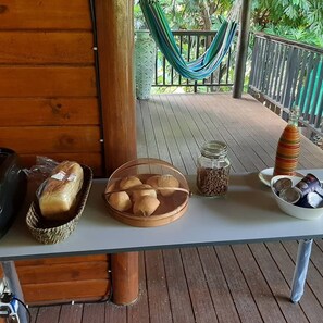 A wooden table with a loaf of bread, a basket of cookies, a jar of cereal, a bottle of syrup, and a bowl of nuts on a wooden deck with a hammock in the background. - Gecko Villa (Praslin Island)