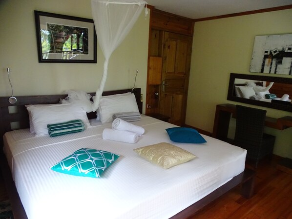 A neatly made bed with pillows and towels, a wooden door, a framed picture on the wall, and a small table with a chair. - Gecko Villa (Praslin Island)