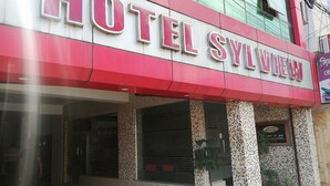 Ett hotell som heter ”HOTEL SYLVIA” med en röd och vit fasad, stora glasfönster och krukväxter vid entrén.