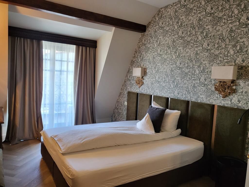 Habitación de hotel con una cama grande, cabecera con estampado floral y vistas al exterior a través de una ventana con cortinas.