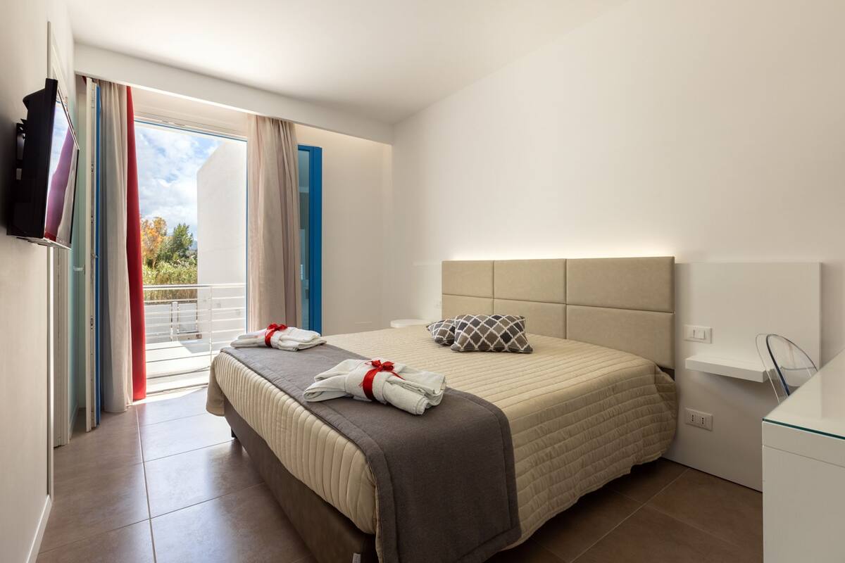 A modern hotel room with a large bed, a flat-screen TV, a glass table, and a balcony with curtains.