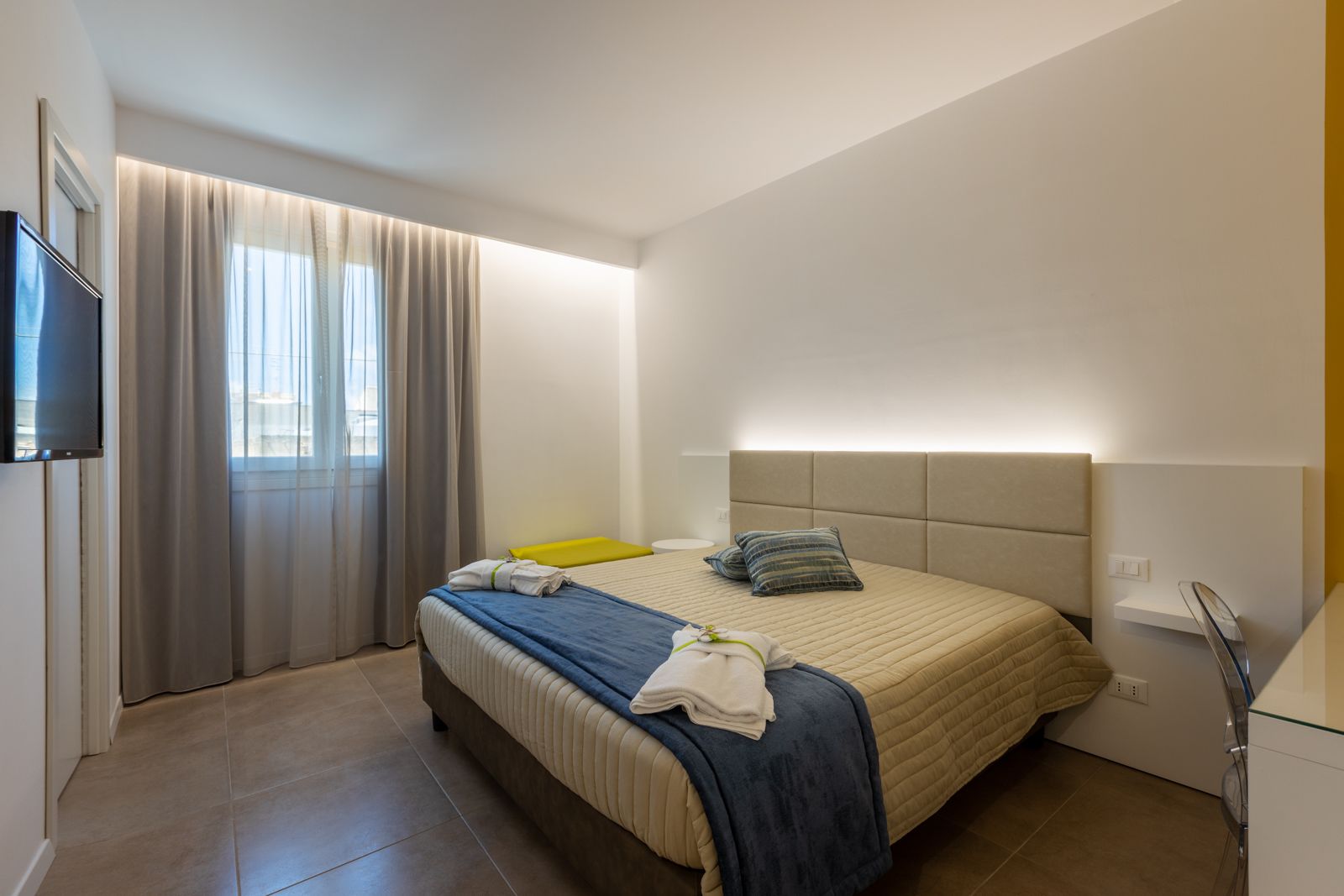 Ein modernes Hotelzimmer mit Bett, Fernseher, Fenster mit Vorhängen und Nachttisch.