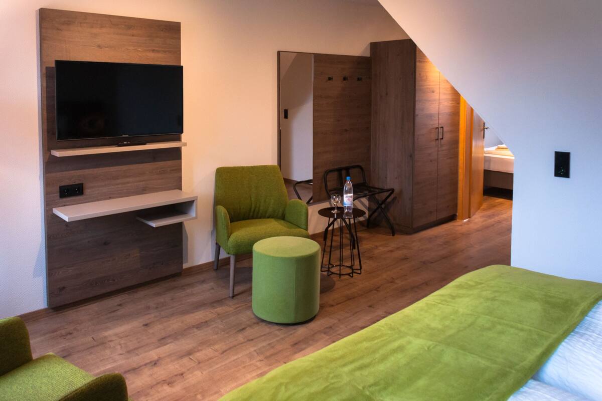 Una habitación de hotel moderna con un televisor de pantalla plana montado en una pared de madera, un sillón verde, un banquillo redondo pequeño, una mesa con una botella y vasos, y una cama con una manta verde.