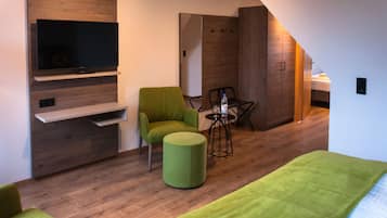 Une chambre d’hôtel moderne avec un téléviseur à écran plat fixé à un mur en bois, un fauteuil vert, un petit pouf rond, une table avec une bouteille et des verres, et un lit recouvert d’une couverture verte.