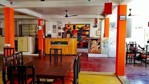 A dining area with a wooden table and chairs, a kitchen in the background, and colorful artwork on the walls. - Rajalakshmi Guest House (Tirukalukundram)