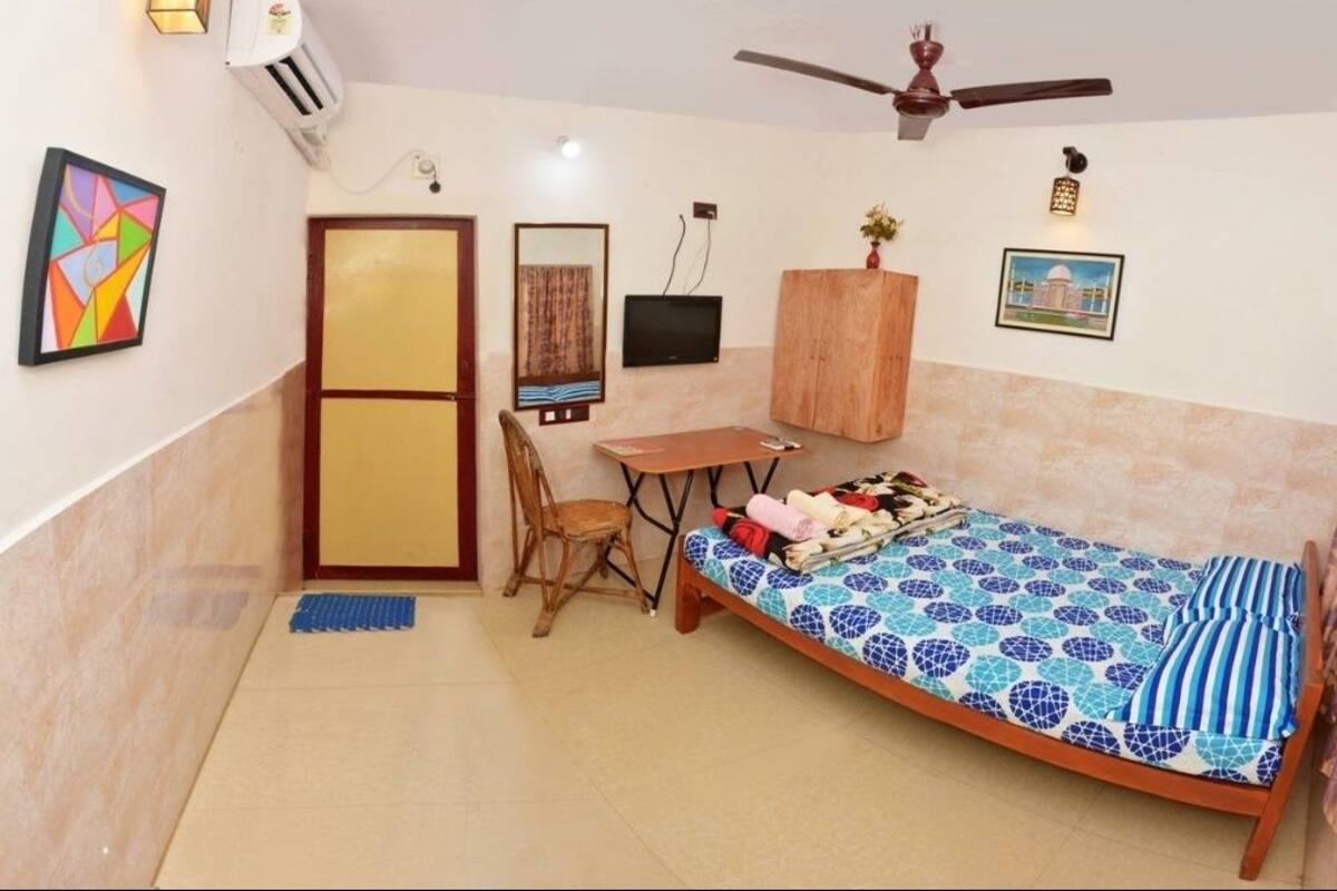 Een kleine kamer met een bed, een tafeltje, een stoel, een televisie en een plafondventilator.