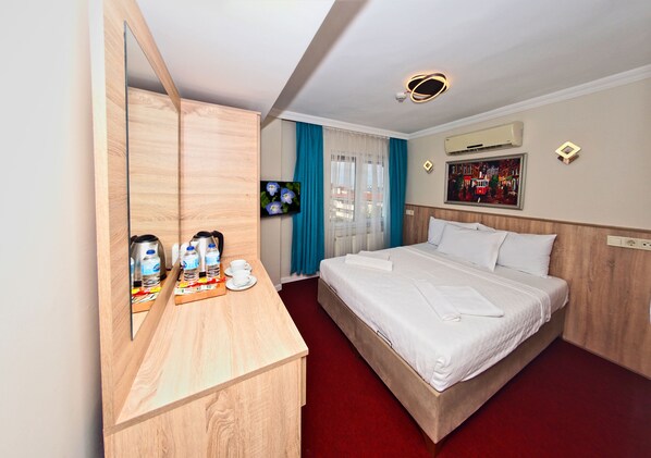 A hotel room with a wooden desk, a bed with white linens, a large mirror, a ceiling light, and a window with blue curtains.