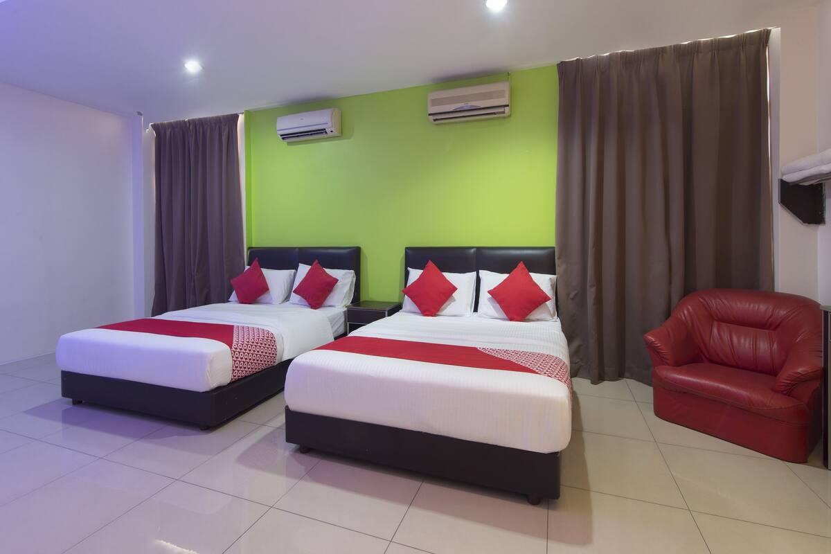 Habitación de hotel con dos camas, ropa de cama roja y blanca, un sillón de cuero rojo y un equipo de aire acondicionado en la pared.