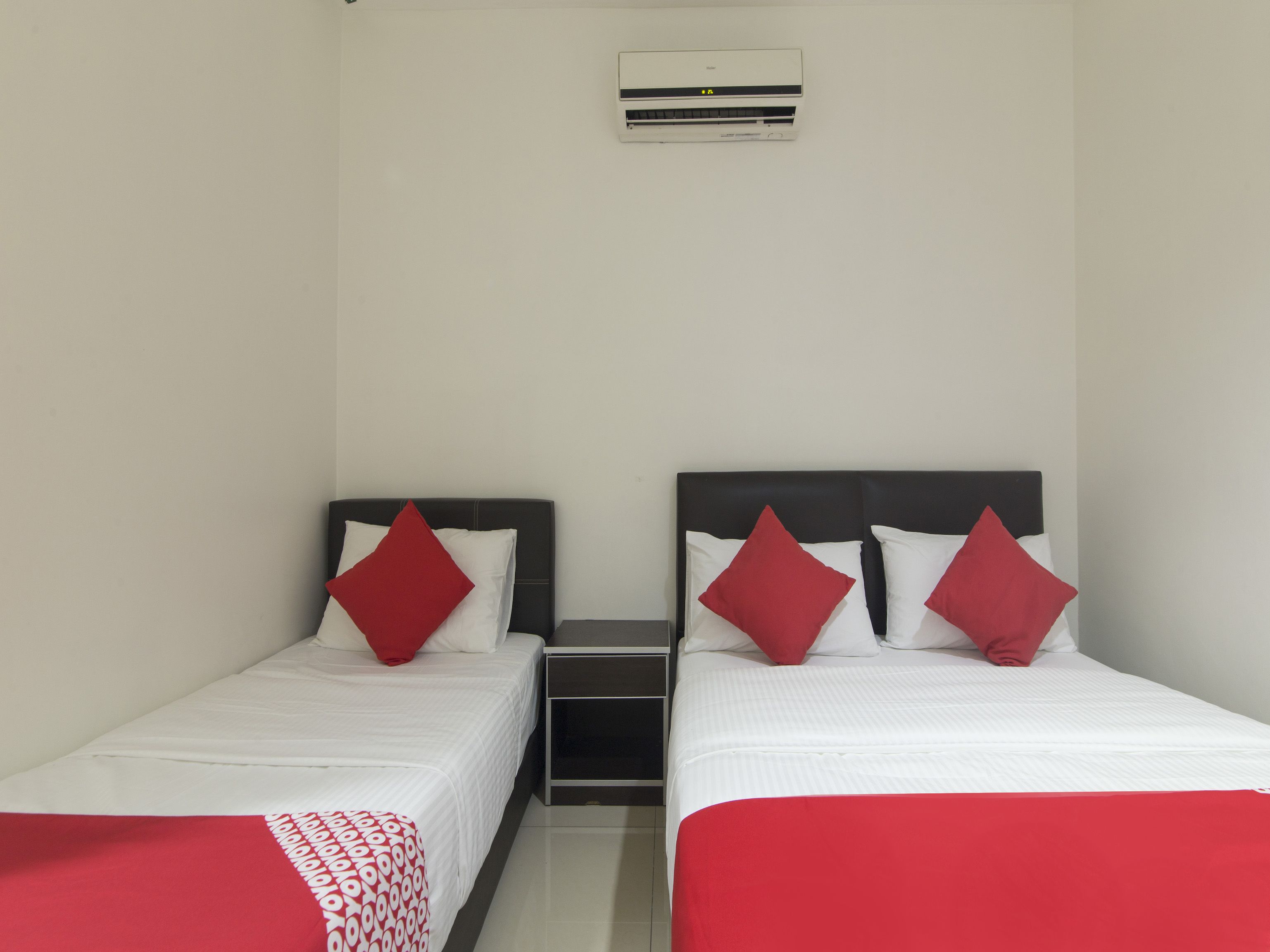 A hotel room with two single beds, each with a red and white pillow, and a small bedside table.