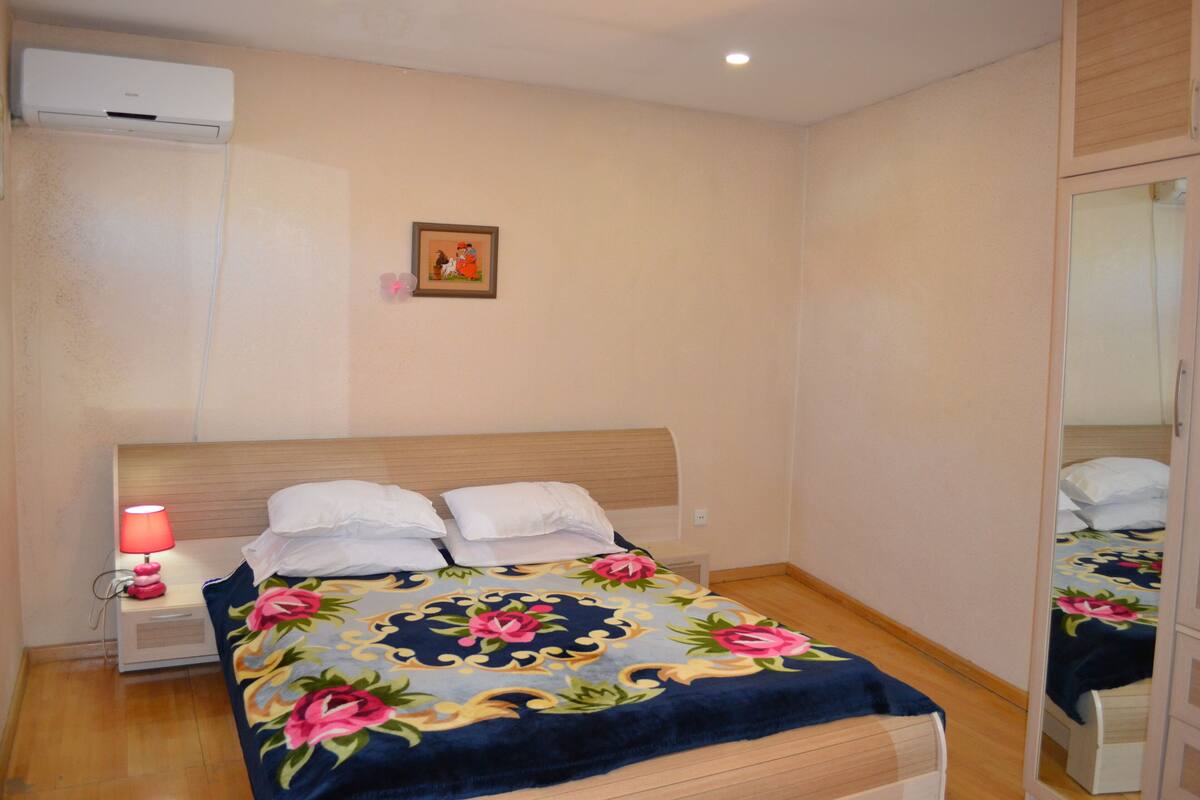 A bedroom with a bed, a nightstand with a lamp, a wall-mounted air conditioner, and a framed picture.