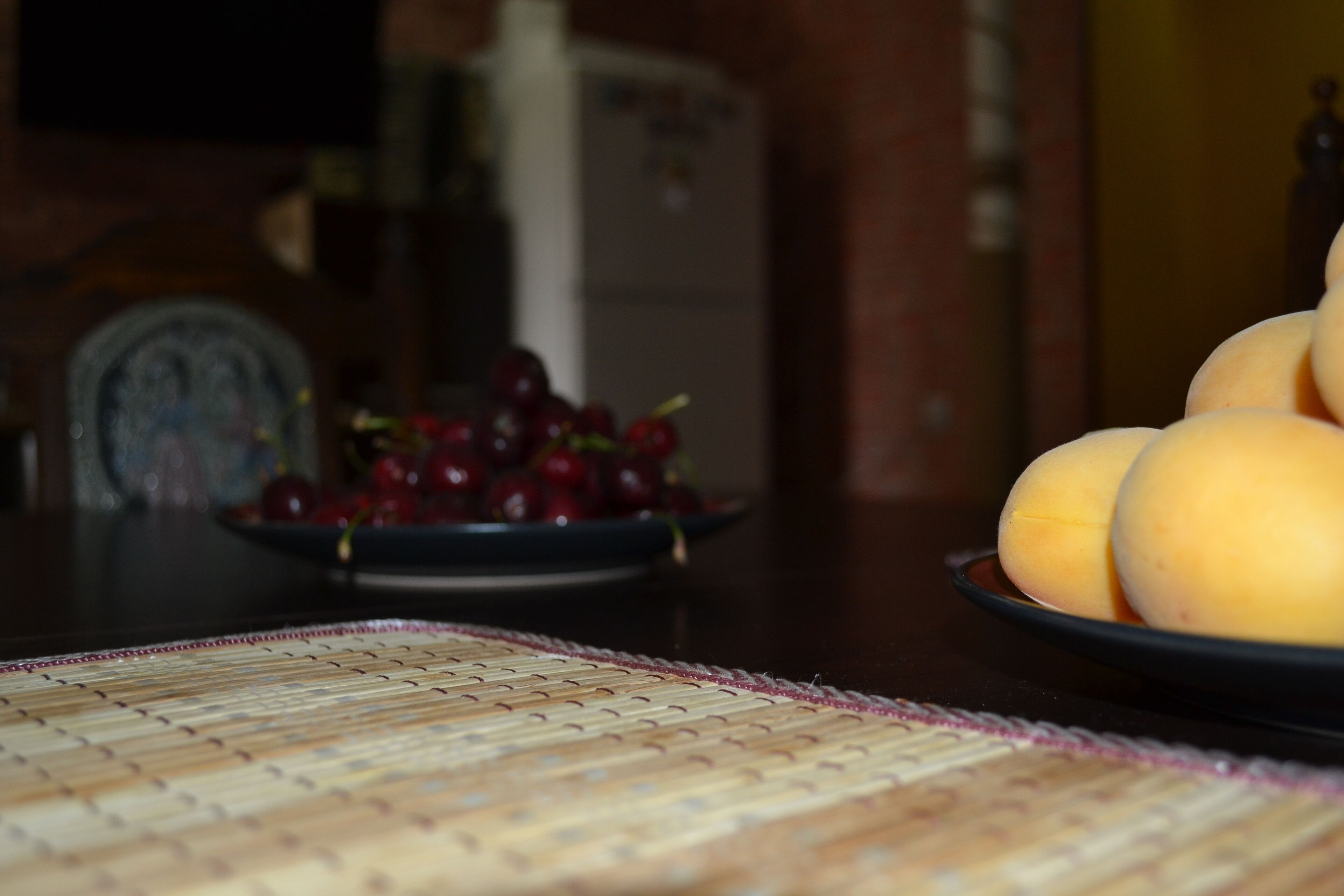 A table with a woven placemat, a plate of yellow fruits, and a bowl of red fruits.