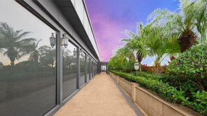 A corridor with glass walls, a tiled floor, and a row of palm trees and shrubs. - Shenzhen Kaijia Hotel (Shenzhen)