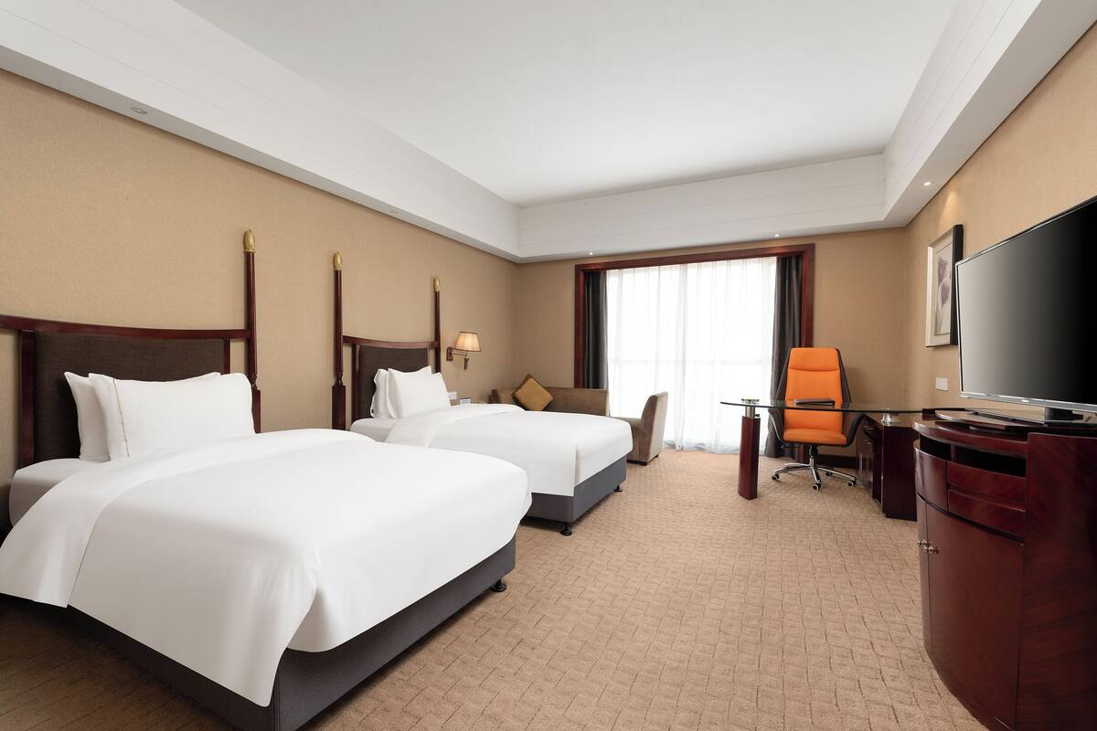 Une chambre d’hôtel avec deux lits, un bureau, une chaise, une télévision et une grande fenêtre.