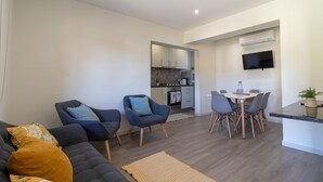 A modern living room with a sofa, two armchairs, a dining table with chairs, a kitchen in the background, and a flat-screen TV mounted on the wall.