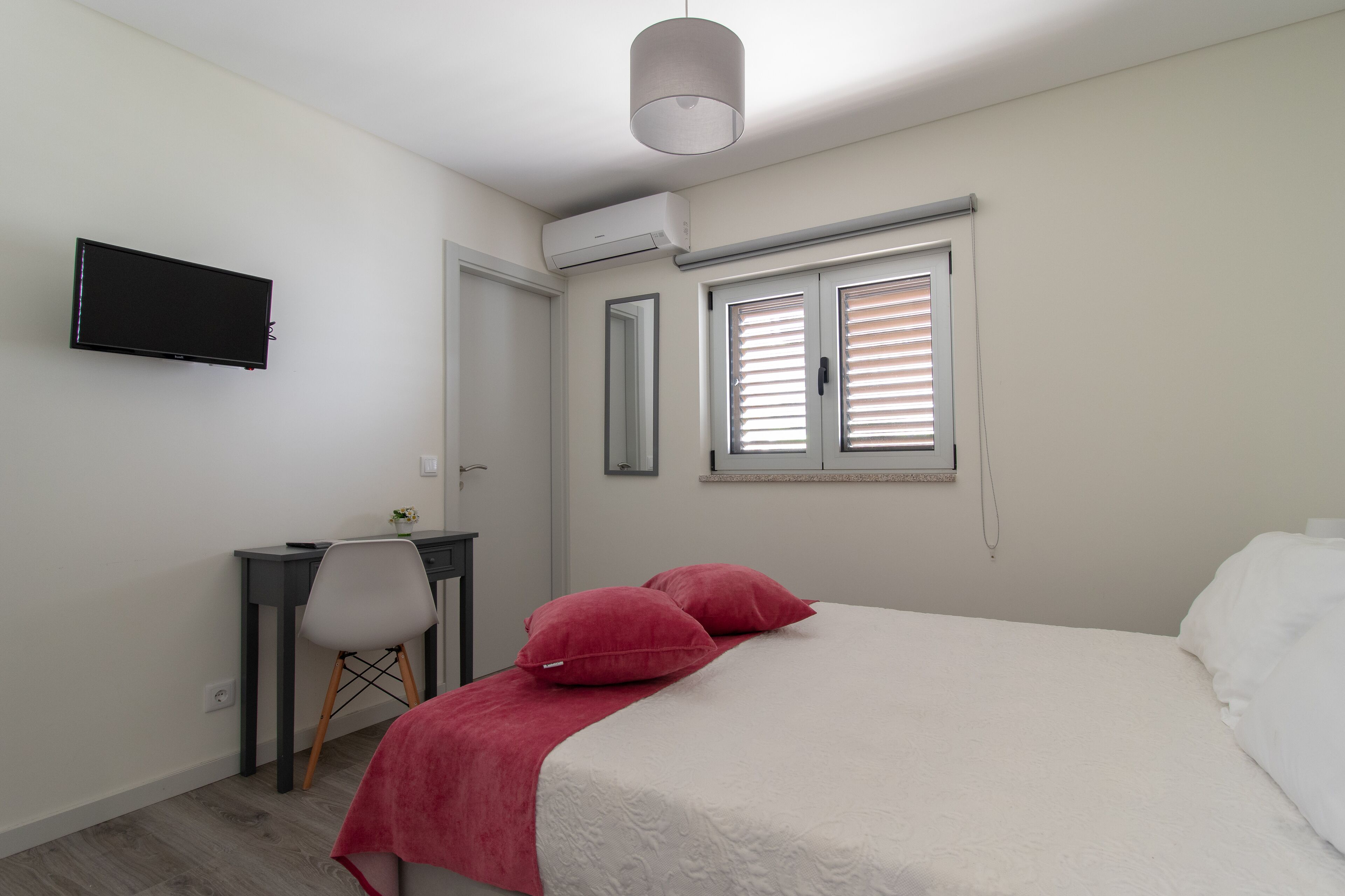 A bedroom with a bed, a desk, a chair, a television, and a window with blinds.