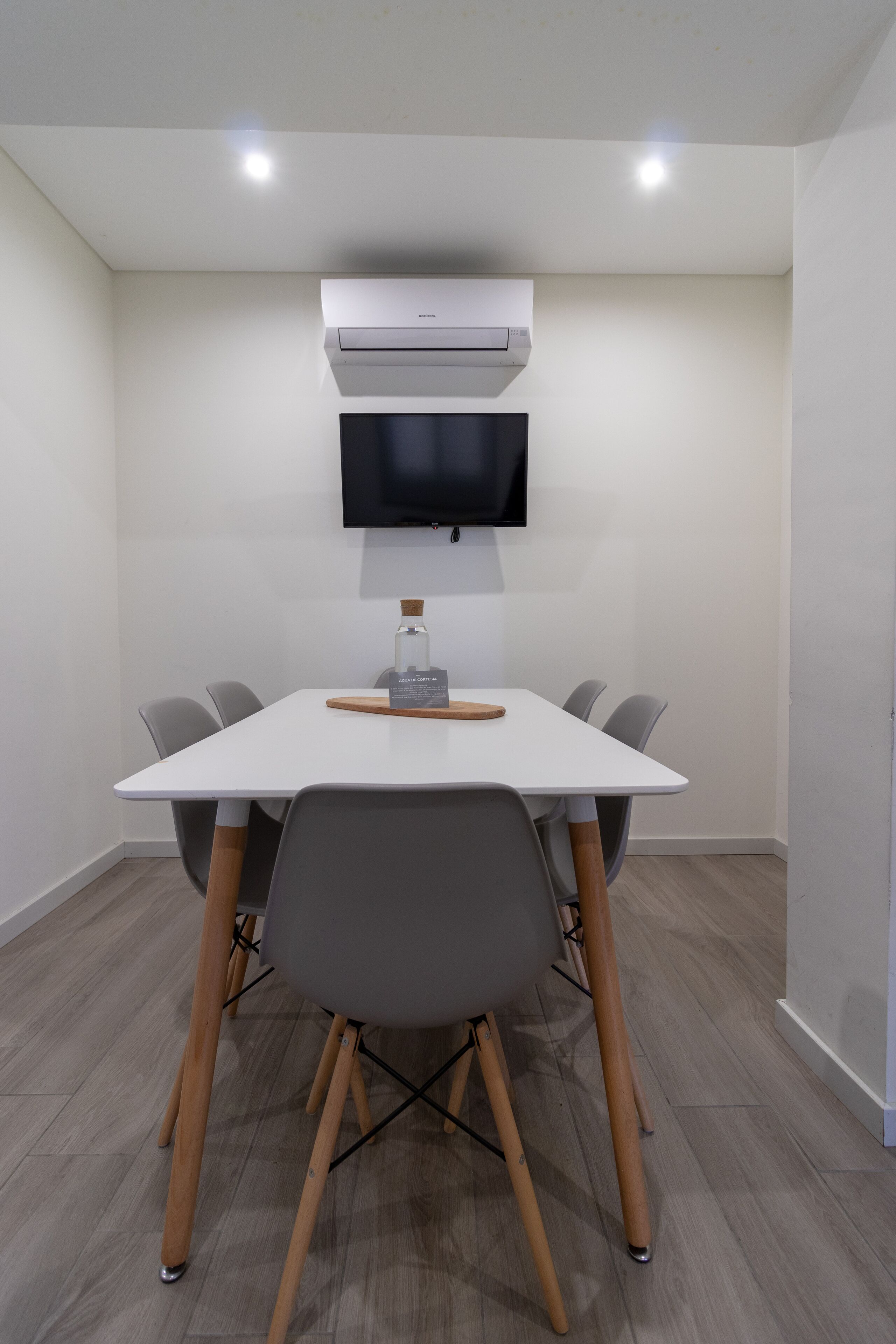 Una sala de reuniones moderna con una mesa blanca, sillas grises y una televisión montada.