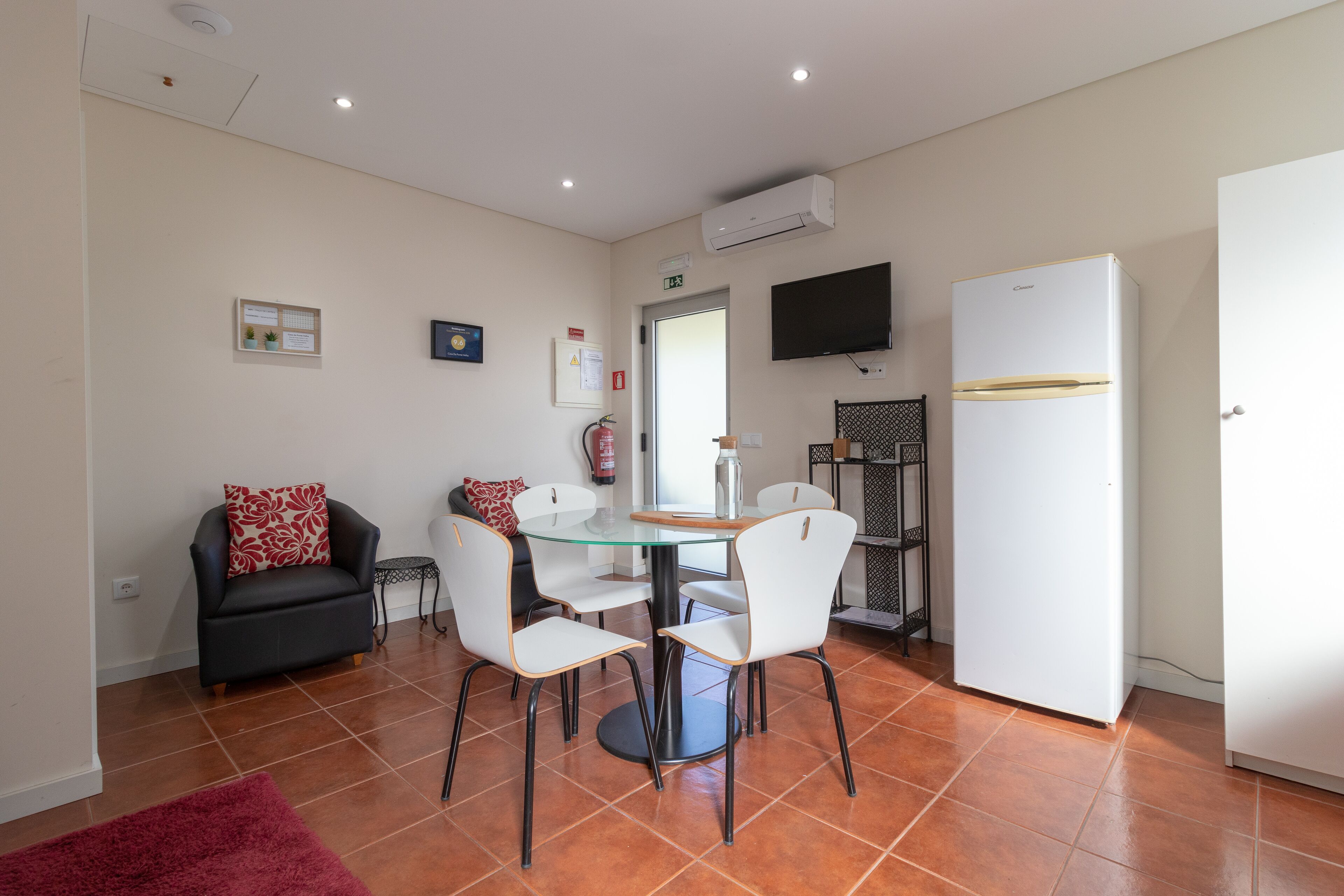 A compact living space with a dining area featuring a glass table and white chairs, a black sofa with red cushions, a white refrigerator, and a mounted TV.