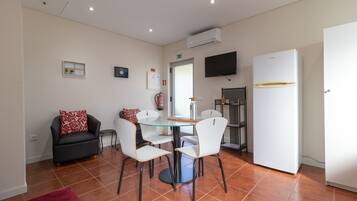 A compact living space with a dining area featuring a glass table and white chairs, a black sofa with red cushions, a white refrigerator, and a mounted TV.