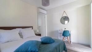 A bedroom with a bed, pillows, a desk, a chair, a mirror, and an air conditioning unit.