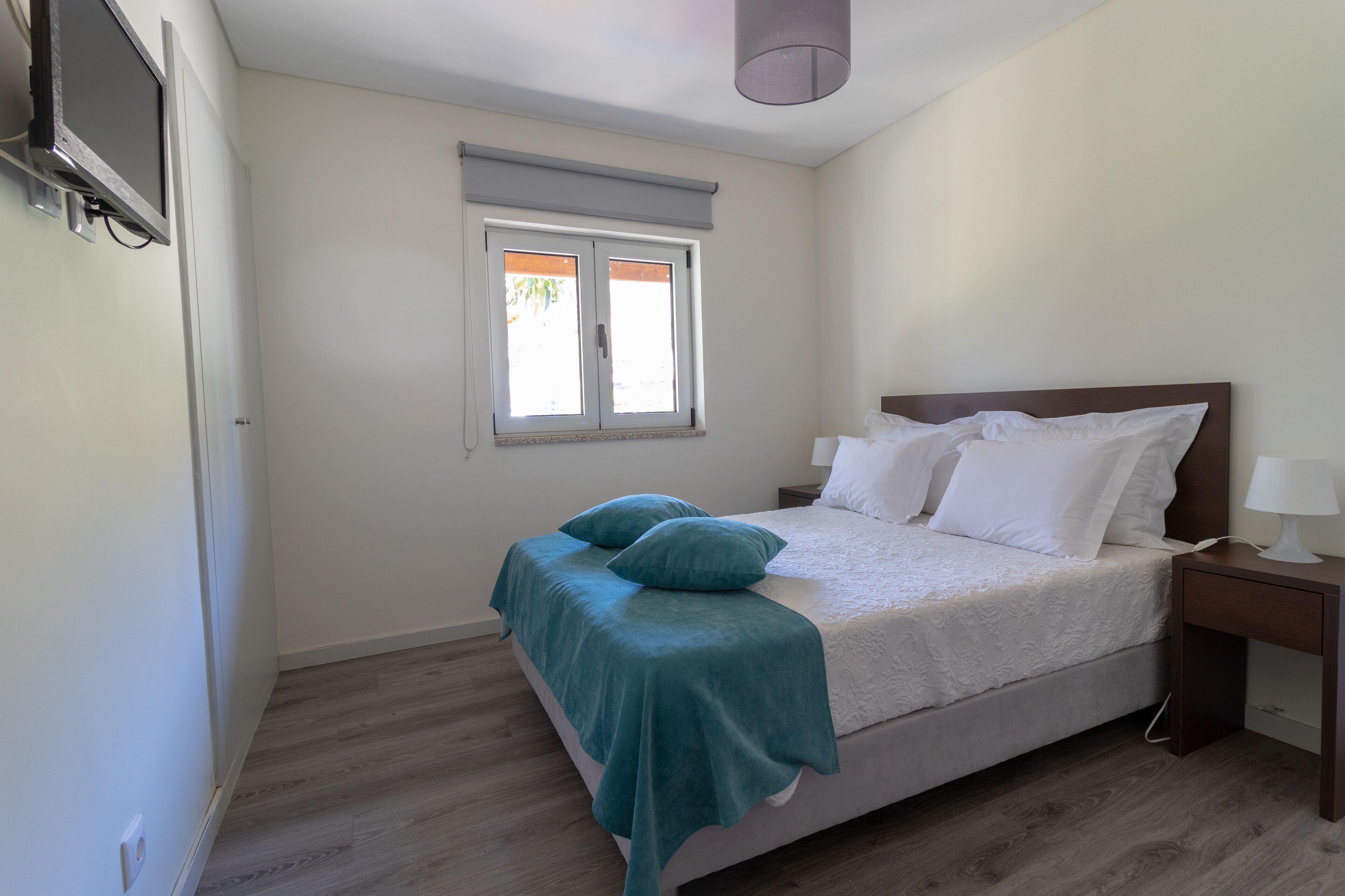 Een slaapkamer met een bed, een raam, een nachtkastje en een tv aan de muur.