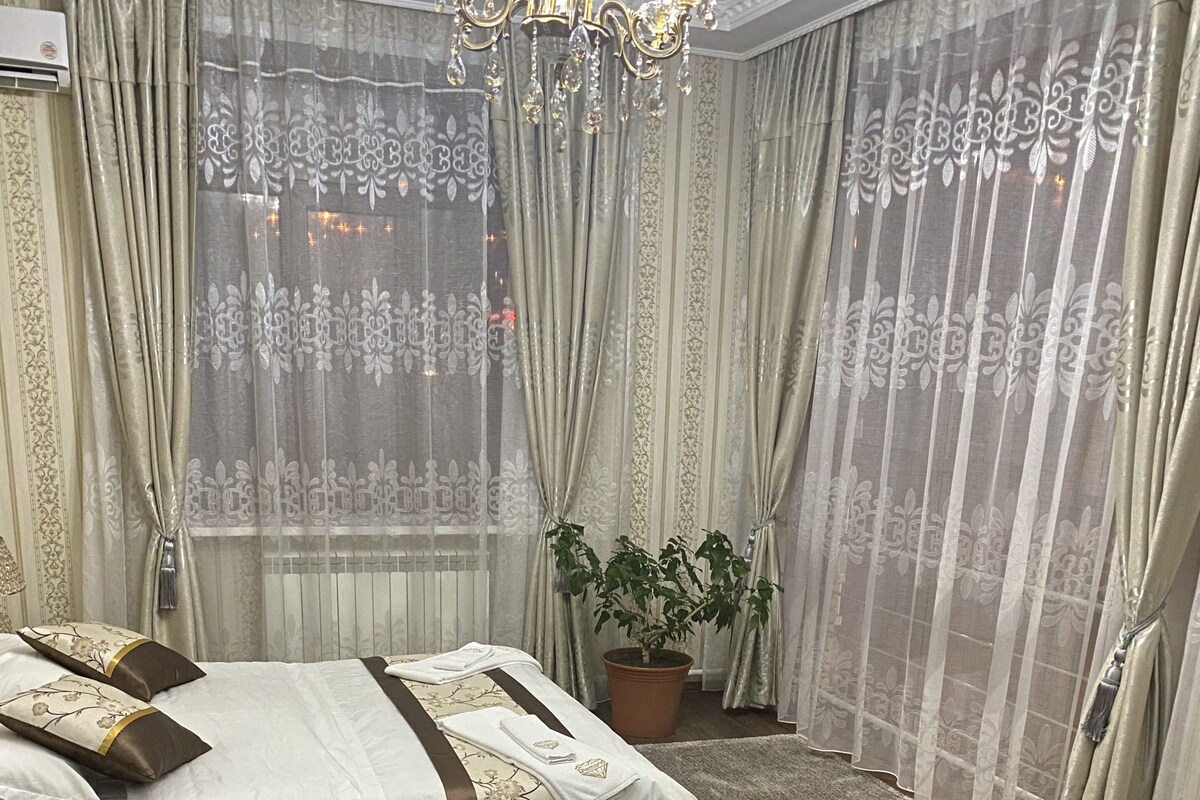 Un dormitorio con cama, cortinas, una araña y una planta en maceta, además de vistas al exterior.