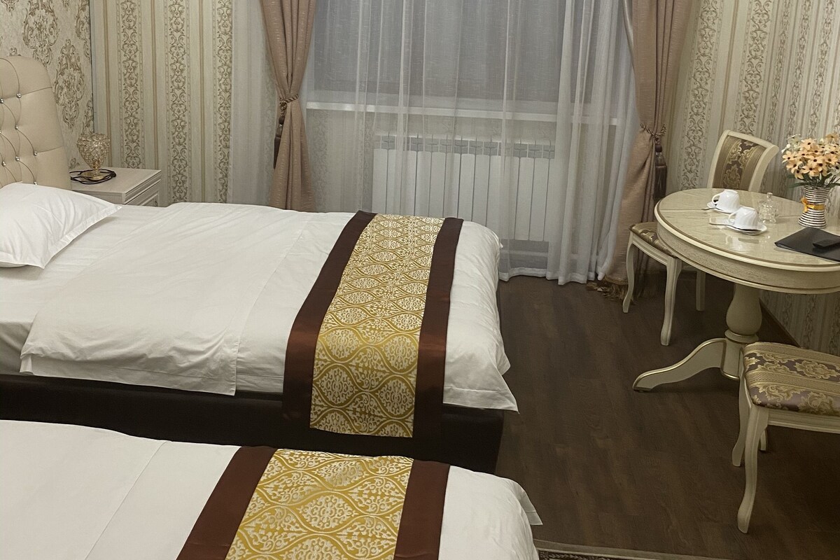 Habitación de hotel con dos camas, un candelabro, una mesa redonda con sillas y vistas a través de cortinas traslúcidas.
