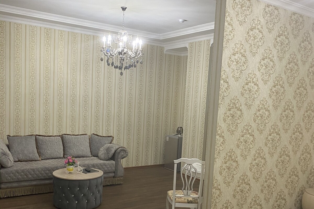Un salón amplio con un sofá gris, un otoman redondos y una silla blanca. La habitación cuenta con papel tapiz a rayas, una araña y una alfombra grande.