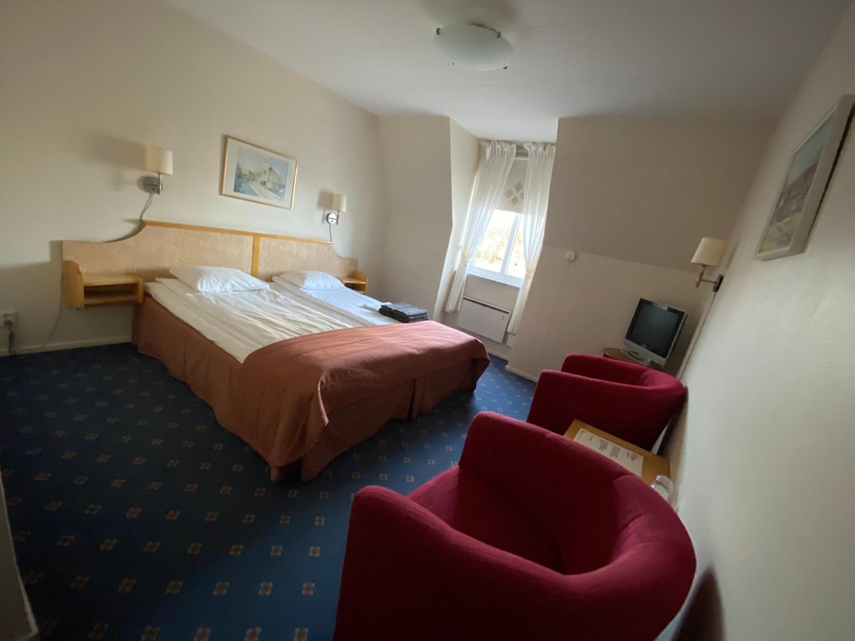 A hotel room with a double bed, a TV, a desk, and two red armchairs.