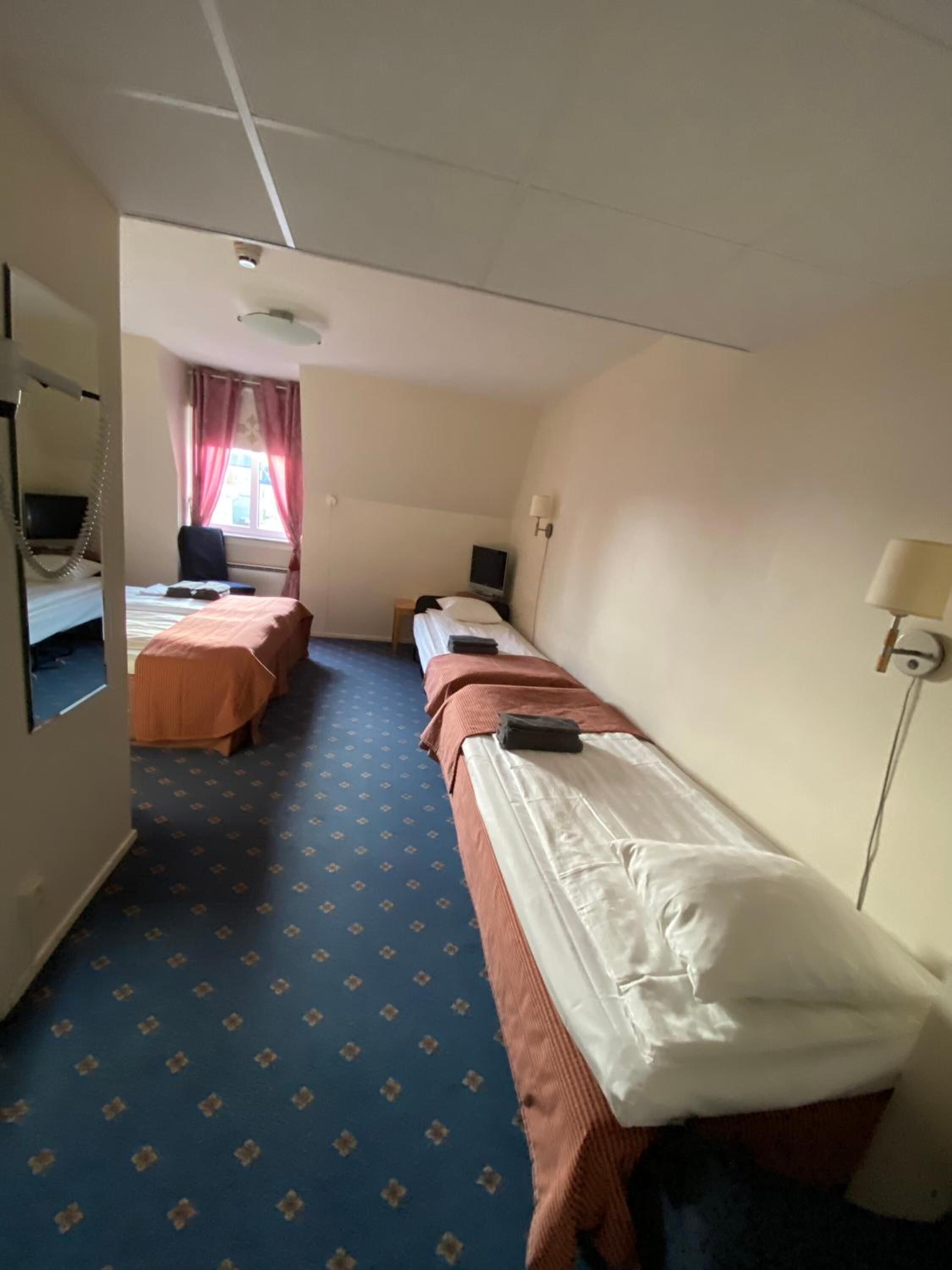 A hotel room with two single beds, a desk with a computer, a chair, a window with curtains, and a lamp.