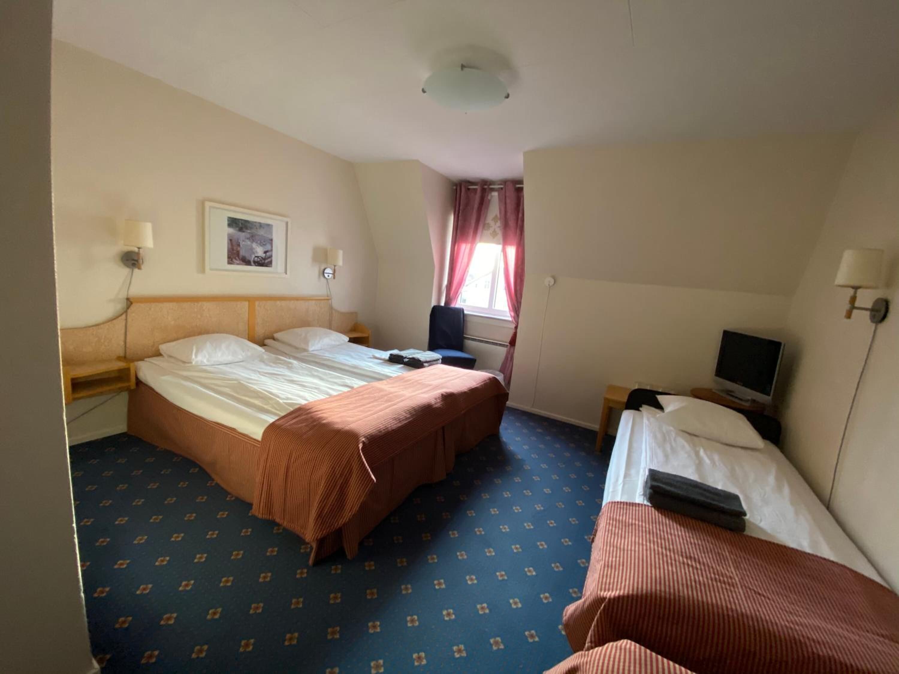 Ett hotellrum med två sängar, ett skrivbord med en dator, en TV och ett fönster med gardiner.