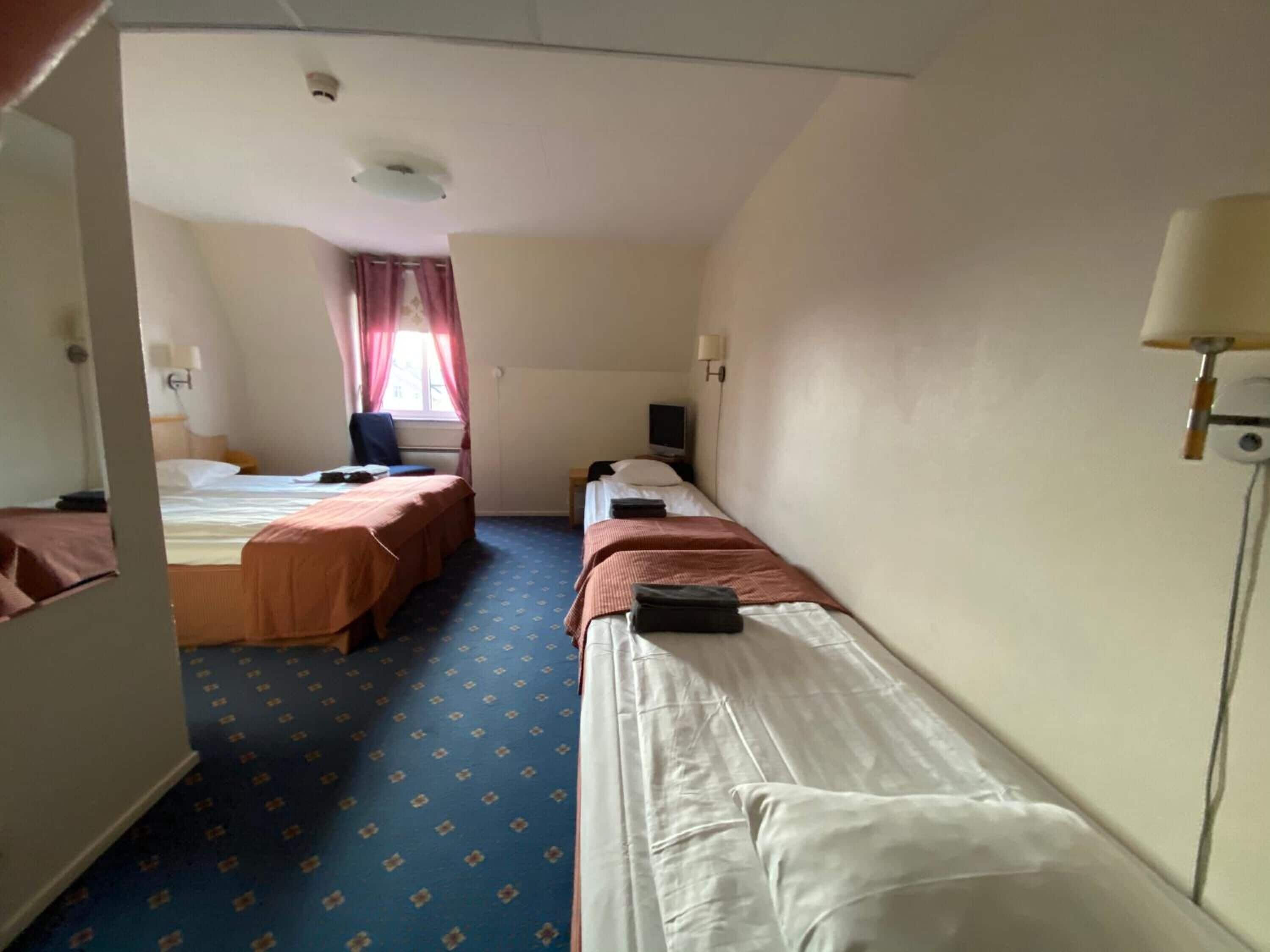 A hotel room with three single beds, a desk, a chair, a TV, and a window with curtains.