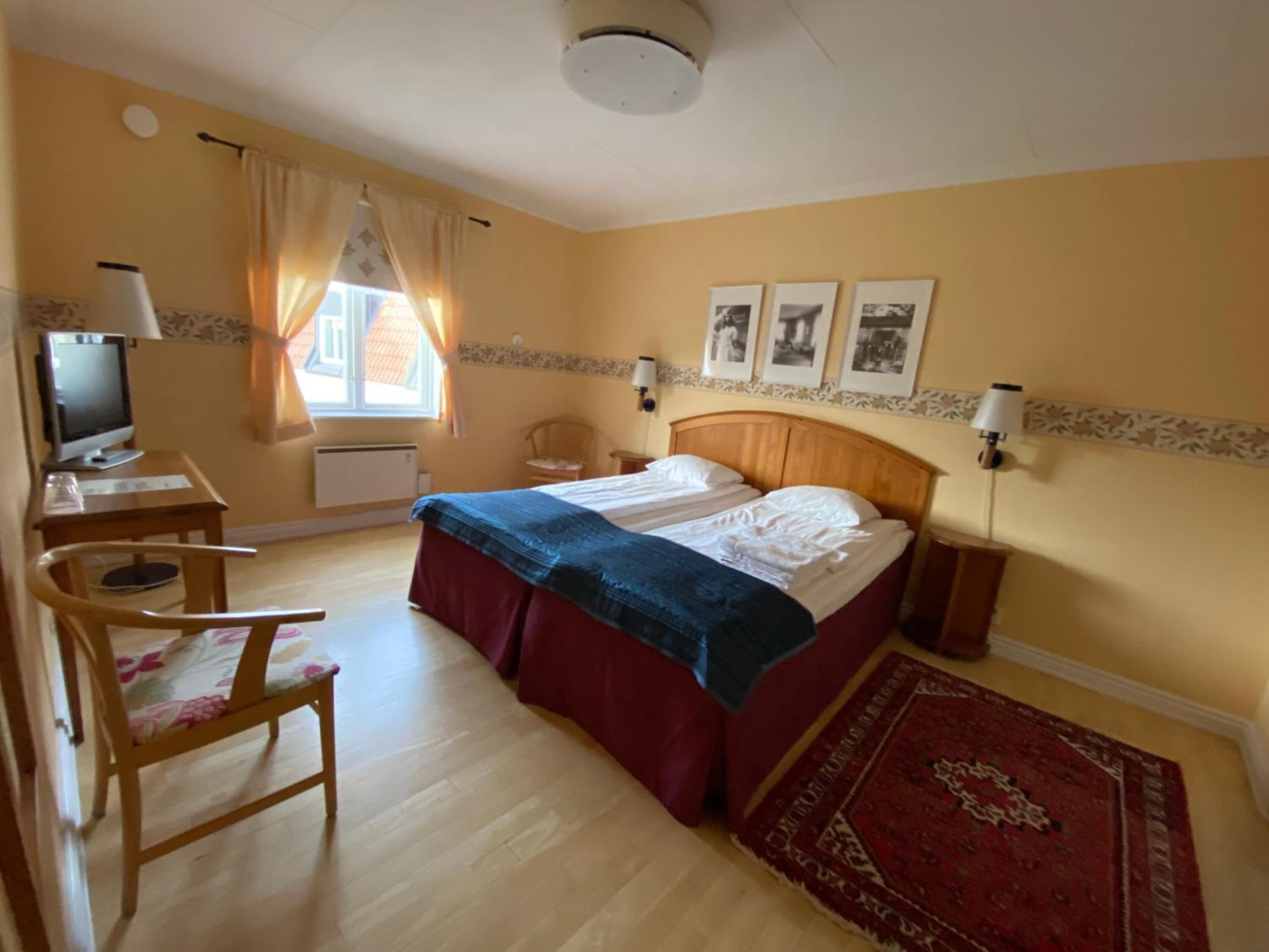A hotel room with a bed, a desk with a computer, a chair, a window with curtains, and a wall with three framed pictures.