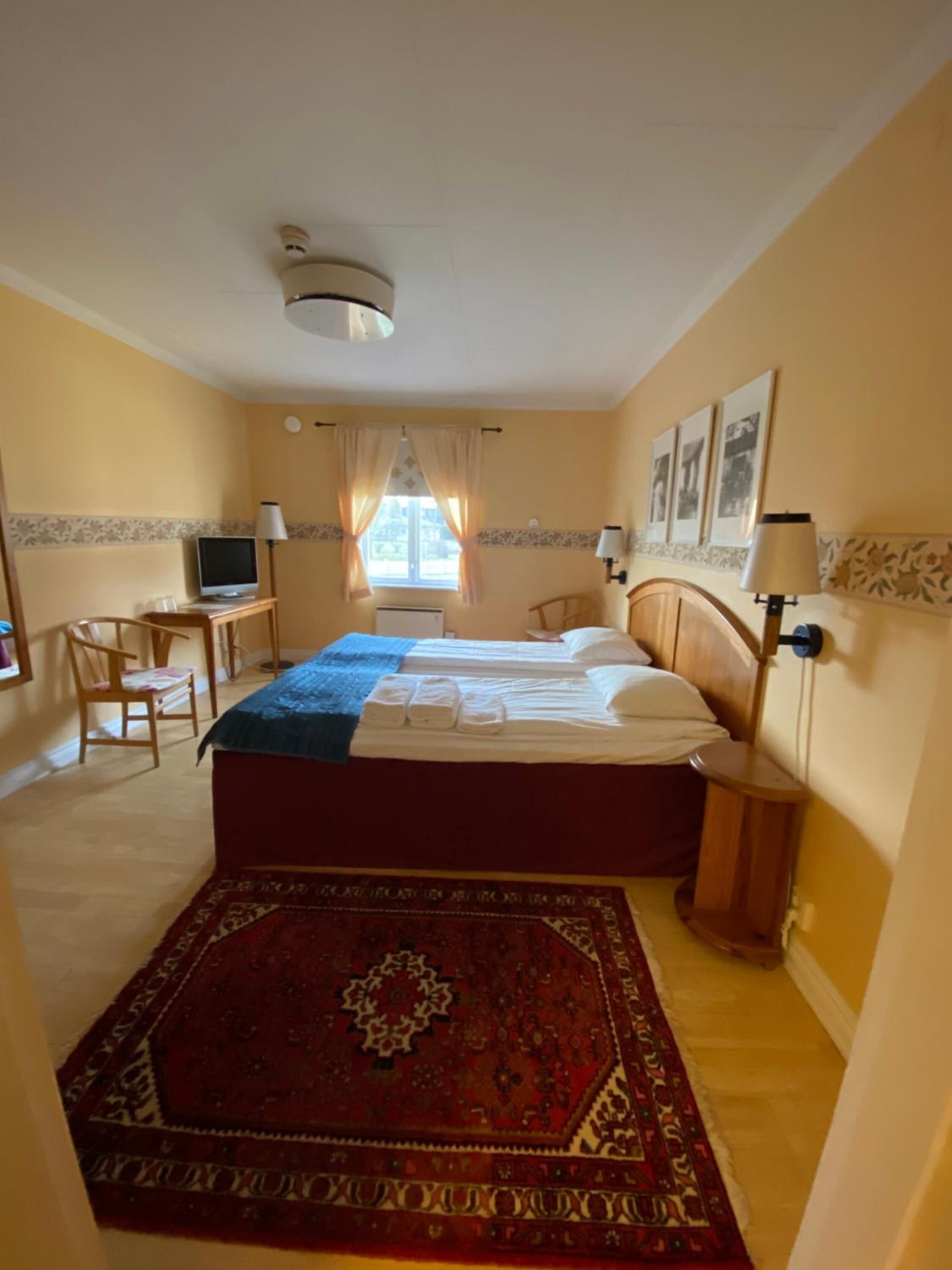 Ein Hotelzimmer mit zwei Betten, einem Schreibtisch mit Computer, einem Fernseher, einem Fenster mit Vorhängen und einem gemusterten Teppich.