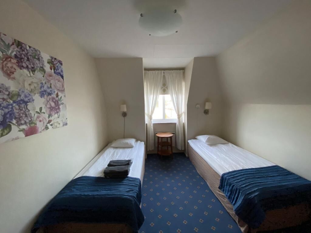 A small, narrow room with two single beds, a small table, and a window with curtains.