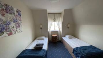 A small, narrow room with two single beds, a small table, and a window with curtains.