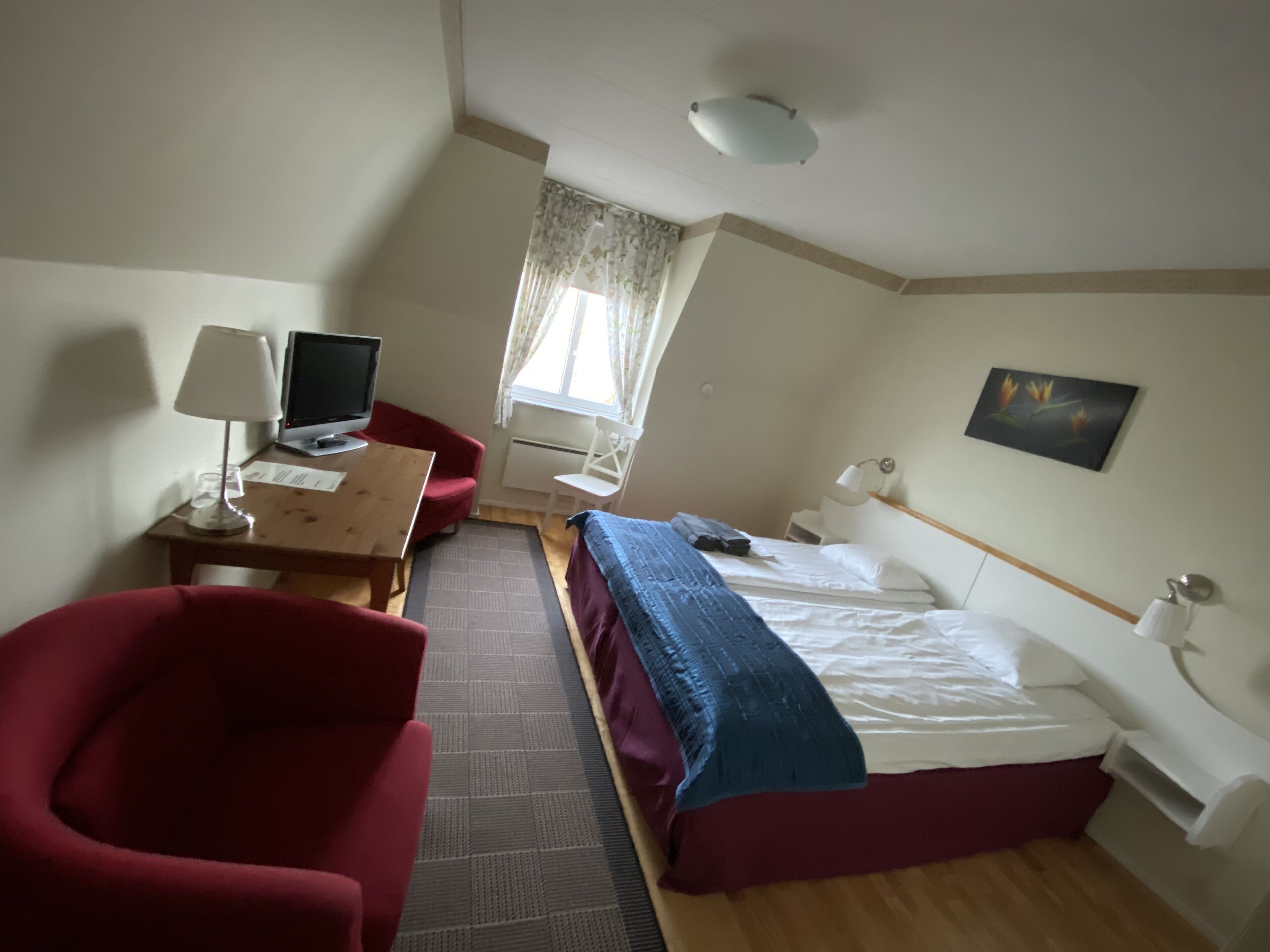 A hotel room with a bed, a desk with a computer, a chair, a lamp, and a window with curtains.