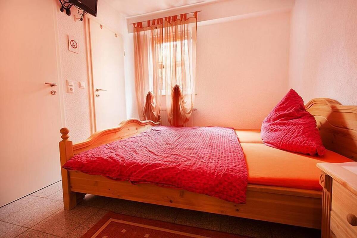 A single bedroom with a wooden bed, a red blanket, and a window with sheer curtains.