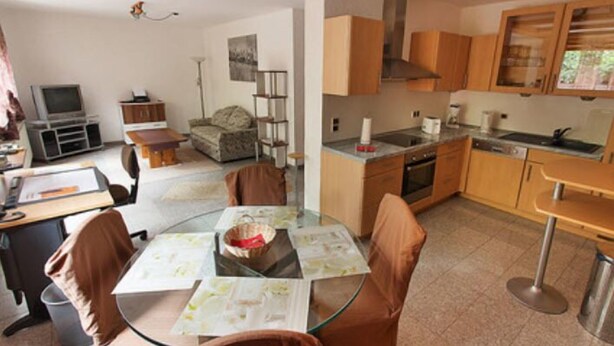 A modern kitchen with wooden cabinets, a dining area with a glass table and brown chairs, and a living area with a sofa and TV. - Gästehaus Walter (Offenbach an der Queich)