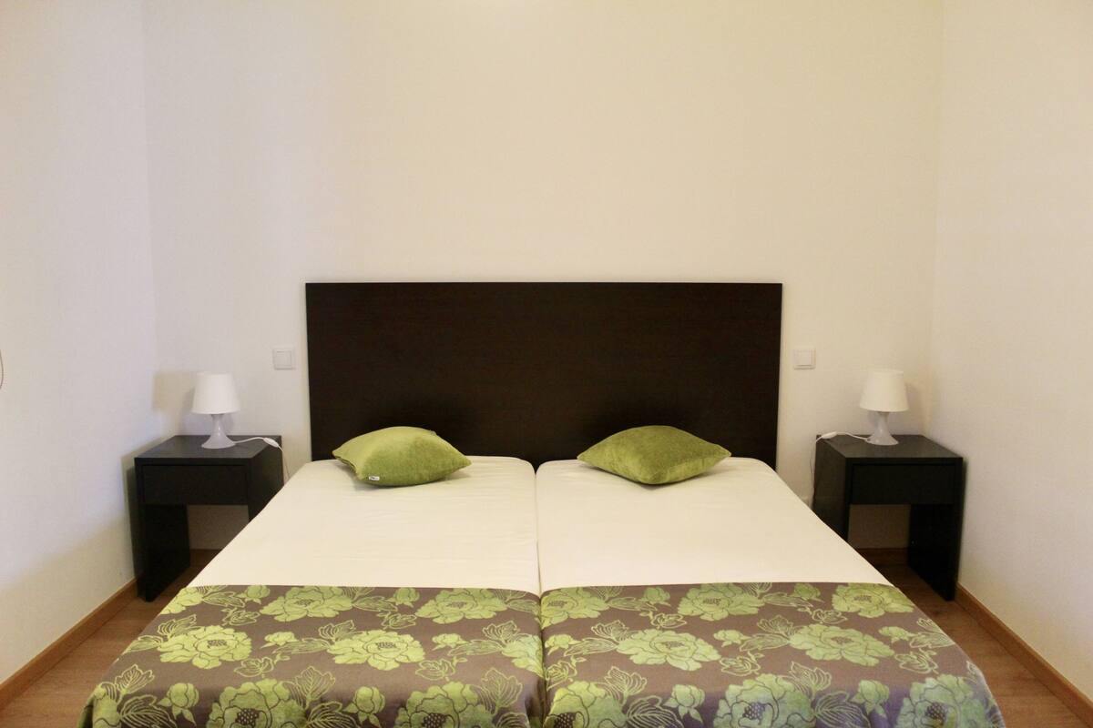 A double bed with a dark headboard, two green pillows, and a floral-patterned bedspread. There are two bedside tables with lamps.